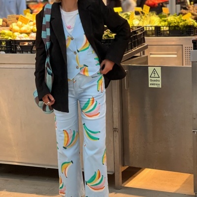 Mulher com fato azul claro de bananas, blazer preto e óculos de sol em mercado com legumes ao fundo