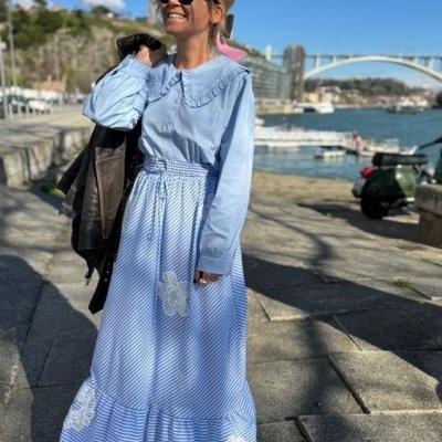 Mulher a usar vestido longo azul claro com riscas brancas e bordados florais, óculos escuros, casaco de cabedal preto ao ombro, ao ar livre junto a um rio e uma ponte