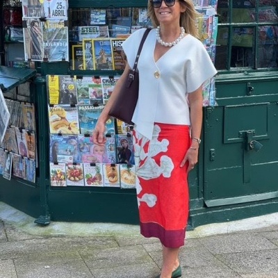 Mulher com blusa branca assimétrica, saia vermelha com padrão branco, sapatos verdes, colar de pérolas e óculos de sol em frente a banca de jornais