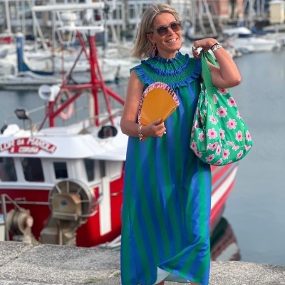 Mulher com vestido de riscas azuis e verdes, leque amarelo e mala verde com flores, em cais com barcos.