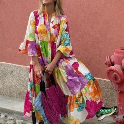 Mulher com vestido floral colorido, mala rosa com franjas e ténis coloridos encostada a parede rosa