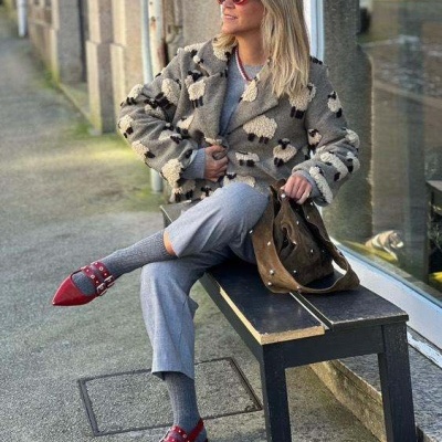 Mulher sentada num banco de madeira com casaco de pelo camuflado, calças cinza, sapatos vermelhos e mala castanha