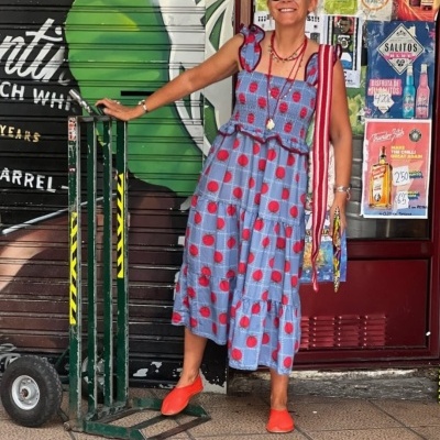 Mulher com vestido azul com bolas vermelhas, chapéu de palha e sapatos vermelhos, junto a carrinho de carga e cartazes