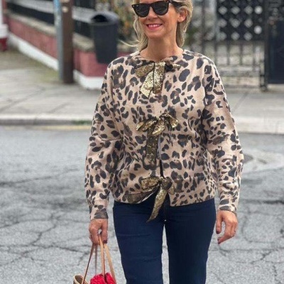 Mulher com cardigan de leopardo e detalhes dourados, calças escuras, óculos de sol, a segurar mala monogramada com lenço vermelho