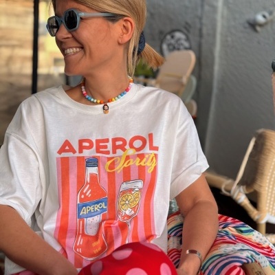 Pessoa com camisola branca APEROL Spritz e calças vermelhas com bolinhas cor-de-rosa sentada