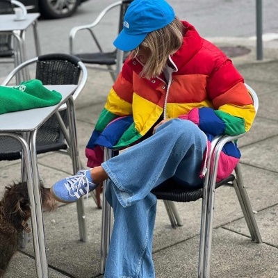 Pessoa sentada com casaco colorido, calças de ganga, ténis azuis e boné azul, com cachorro próximo numa esplanada
