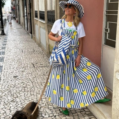 Mulher com saia, chapéu e bolsa às riscas azuis e brancas com limões, t-shirt e sapatos verdes, a passear cão castanho
