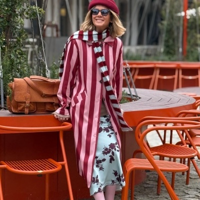 Mulher com casaco riscas rosa e vermelho, saia azul clara com flores, cachecol, boina vermelha e óculos escuros, numa esplanada laranja.