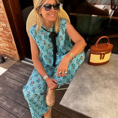 Mulher sorridente com roupa azul floral e óculos escuros sentada em esplanada com bolsa castanha