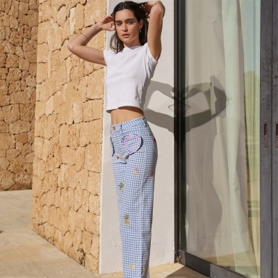 Mulher a usar calças vichy azuis com bordados e t-shirt branca junto a parede de pedra e porta de vidro