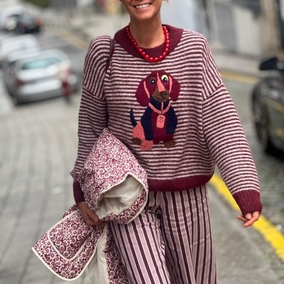 Mulher com camisola riscada e padrão cão, calças listradas, colar vermelho, óculos escuros e chapéu, segurando casaco floral