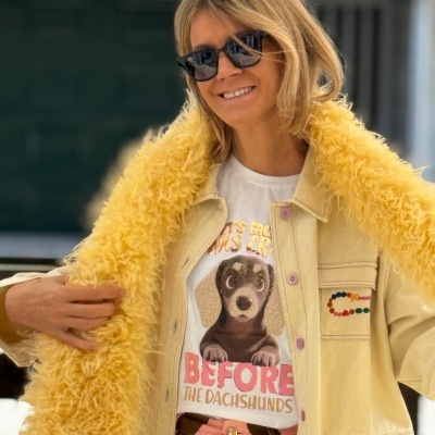 Mulher com casaco amarelo claro com forro amarelo felpudo, t-shirt branca com cão e texto, calças riscas cor de rosa e castanho, óculos escuros pretos