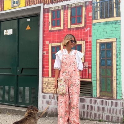 Mulher com macacão floral rosa claro, camisa branca e óculos escuros, com cão marrom em trela frente a parede pintada colorida.
