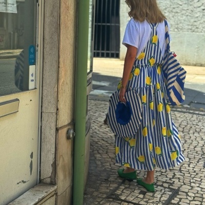 Pessoa com vestido longo às riscas azuis e limões, chapéu e saco combinando, sapatos verdes em rua com mosaico português