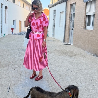 Mulher com conjunto de blusa e saia rosa e vermelha a passear cão de pelo escuro numa rua estreita.