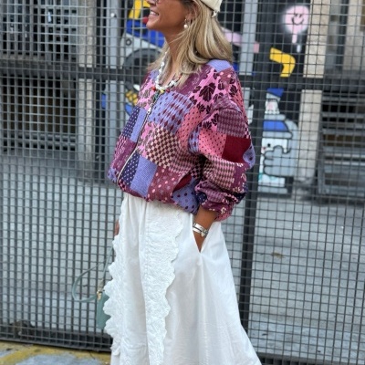 Mulher com casaco patchwork rosa e saia branca em frente a portão metálico com grafite colorido