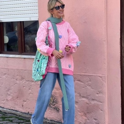 Mulher com camisola rosa, calças largas azuis, ténis e acessórios verdes junto a parede rosa