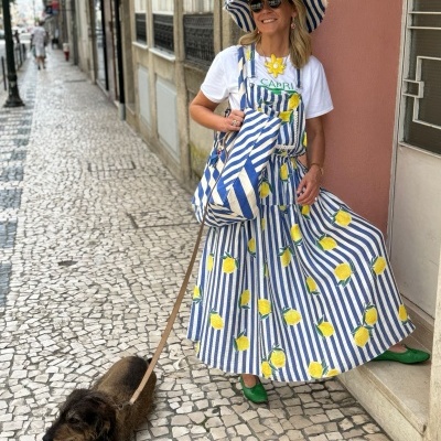 Mulher com vestido azul e branco com limões e chapéu combinando, com cão numa calçada 