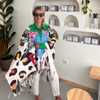 Pashmina colorida com padrão abstrato usada por mulher em ambiente interno com prateleiras e plantas.