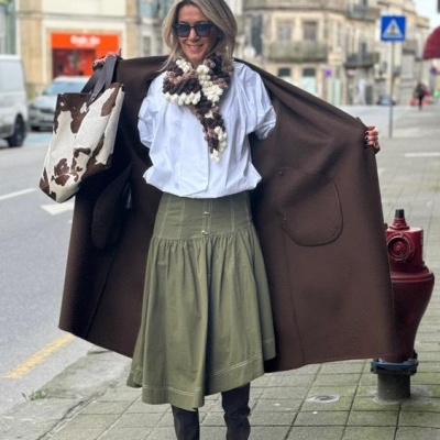 Mulher na rua com casaco castanho longo, camisa branca, saia verde, botas cinzentas, sciarpe de malha e mala padrão pele de vaca