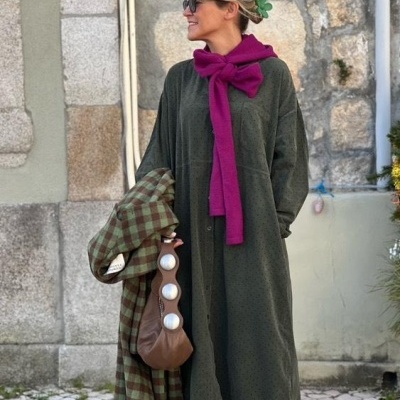 Mulher com vestido verde escuro, echarpe rosa, casaco verde às riscas na mão e bolsa castanha com botões brancos
