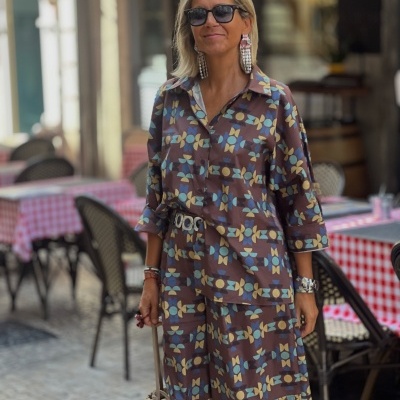 Mulher com conjunto castanho estampado geométrico, brincos grandes, óculos escuros e mala bege