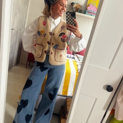 Mulher com colete bege de lã e calças de ganga com corações pretos, tirando selfie num espelho.