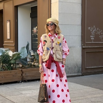 Mulher com vestido rosa de bolinhas vermelhas, colete bege felpudo, chapéu de palha, óculos escuros, sapatos castanhos e bolsa Louis Vuitton numa rua urbana