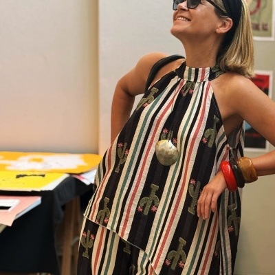 Mulher com vestido de riscas verticais coloridas e bordados de cactos, pulseiras grandes e óculos de sol.