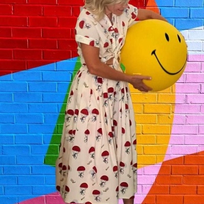 Mulher com vestido creme com padrão de cogumelos segurando balão amarelo sorridente em frente a parede colorida