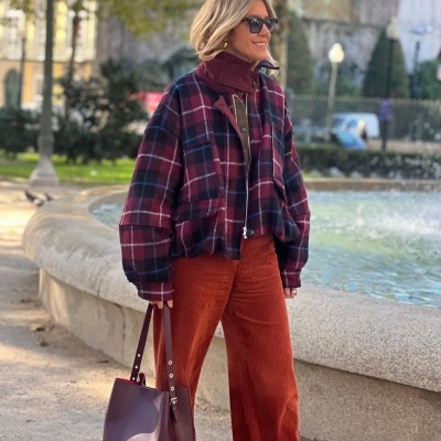 Mulher com casaco xadrez bordeaux e azul escuro com calças ferrugem e bolsa bordeaux junto a fonte