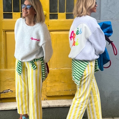 Duas mulheres com calças às riscas amarelas e brancas e camisolas cinza com texto colorido, em frente a porta amarela.