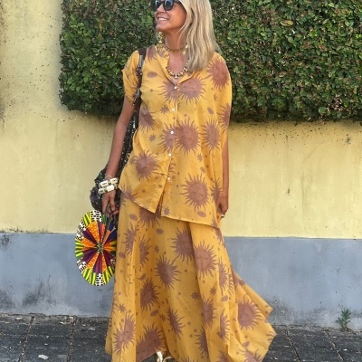 Mulher com conjunto amarelo estampado de flores castanhas, sandálias douradas, óculos escuros e bolsa colorida redonda em rua com parede amarela e arbusto verde.