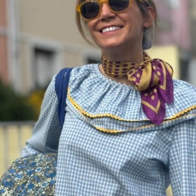 Pessoa com óculos de sol, lenço roxo e amarelo, t-shirt azul xadrez com gola folhada e bolsa azul floral