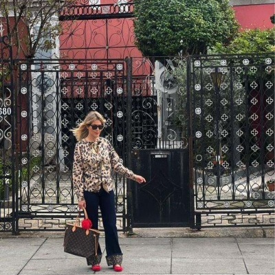 Mulher em blusa animal print, calças pretas e sapatos vermelhos junto a portão de ferro preto decorado.