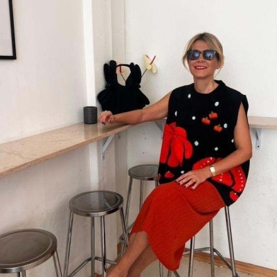 Mulher sentada em banco alto com blusa preta estampada e saia laranja num espaço interior