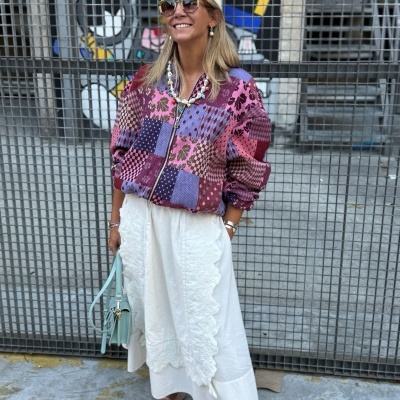 Mulher com blusão colorido patchwork, saia branca rendada, sapatilhas rosa e chapéu branco junto a grade metálica com grafite