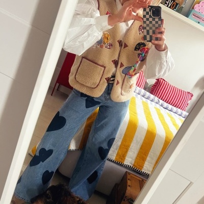 Pessoa a tirar selfie com colete bege felpudo e calças de ganga com corações num quarto com cama às riscas amarelas e brancas.