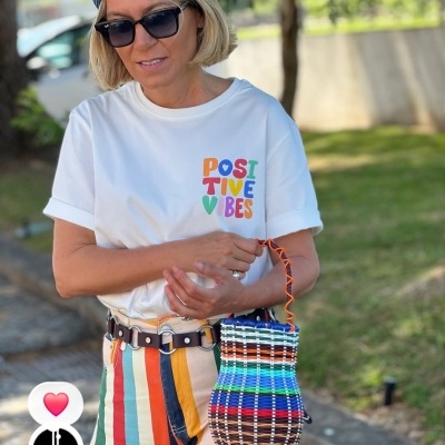 Mulher com t-shirt branca estampada, calças coloridas, chapéu azul, óculos de sol e bolsa tecida multicolorida