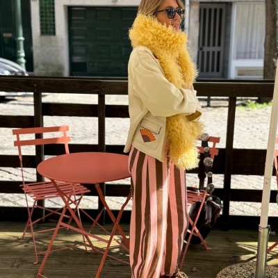 Pessoa com casaco beige, cachecol amarelo, calças às riscas rosa e castanho, ténis castanhos, perto de mesa e cadeiras rosa