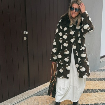 Mulher com casaco felpudo castanho e branco com padrão de ovelhas, vestido branco, sapatos castanhos, óculos escuros e mala castanha num passeio