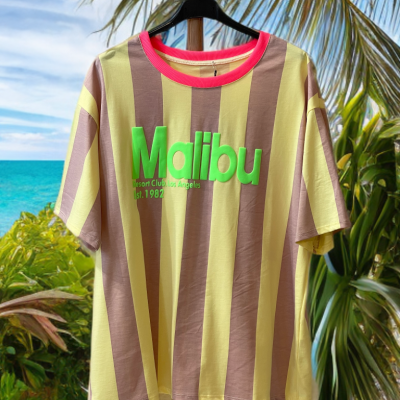 Camisola às riscas castanho e amarelo com texto verde Malibu pendurada em cabide diante de cenário de praia
