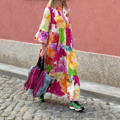 Mulher com vestido floral colorido e mala azul com franjas andando numa rua com parede rosa