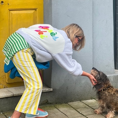 Pessoa com roupa colorida acaricia cão pequeno junto a porta amarela e parede cinza