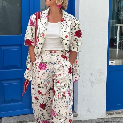 Conjunto de roupa com padrão floral colorido em fundo creme, top branco e sapatos vermelhos, mulher em frente a porta azul.