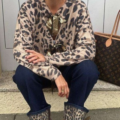 Mulher sentada com suéter animal print bege e castanho, calças azul-escuro com barra animal print e sapatos vermelhos, ao lado mala de couro castanho com monograma.