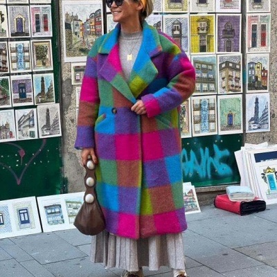 Mulher com casaco colorido e saia plissada numa rua urbana com quadros de arquitectura à venda