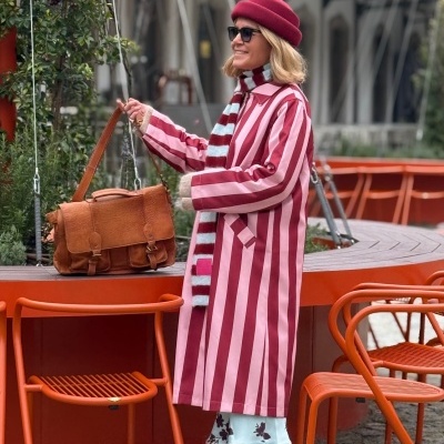 Mulher de casaco riscado rosa e vermelho com boina vermelha e mala de couro em área exterior com cadeiras laranja