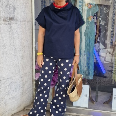 Mulher com camisa preta, calças de bolinhas e mala de palha em frente a loja