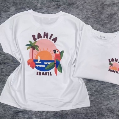 Duas t-shirts brancas com estampa colorida de pôr do sol e papagaio, sobre textura felpuda cinzenta.